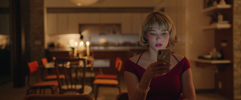A woman with a blonde bob in a red dress looks at her phone in a warm, candlelit dining room