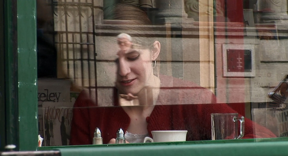 A woman in a red cardigan seen through a cafe window with Manhattan street reflections on the glass